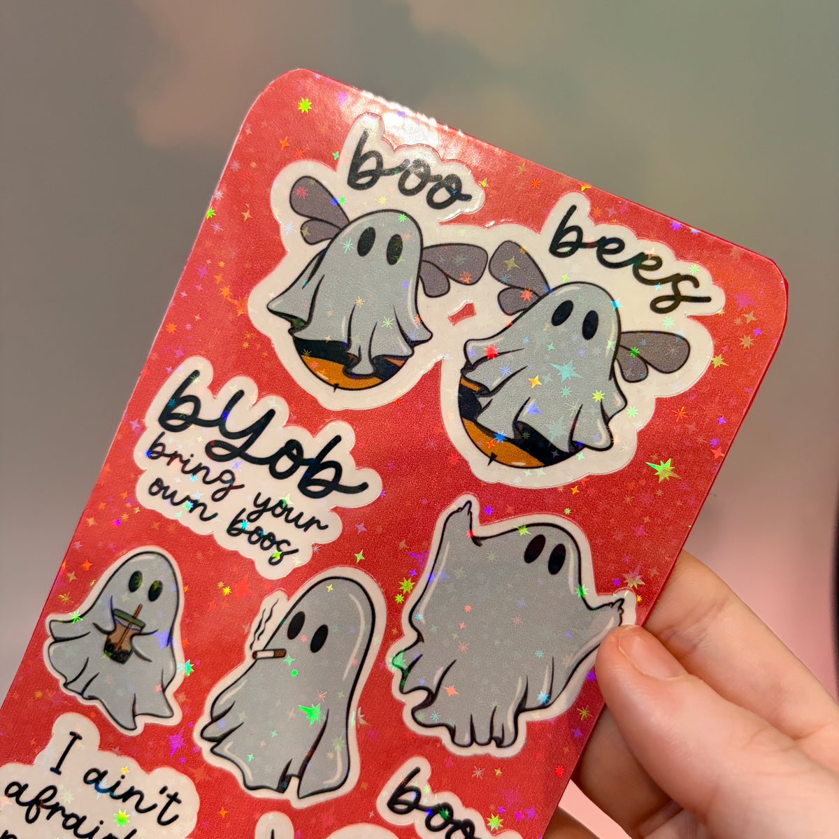 Holographic sticker sheet featuring eight cute ghost vinyl stickers with funny sayings including "Boo Bees," "BYOB Bring Your Own Boos," "I Ain't Afraid of No Ghost," and other snarky spooky designs on a pink glitter background.