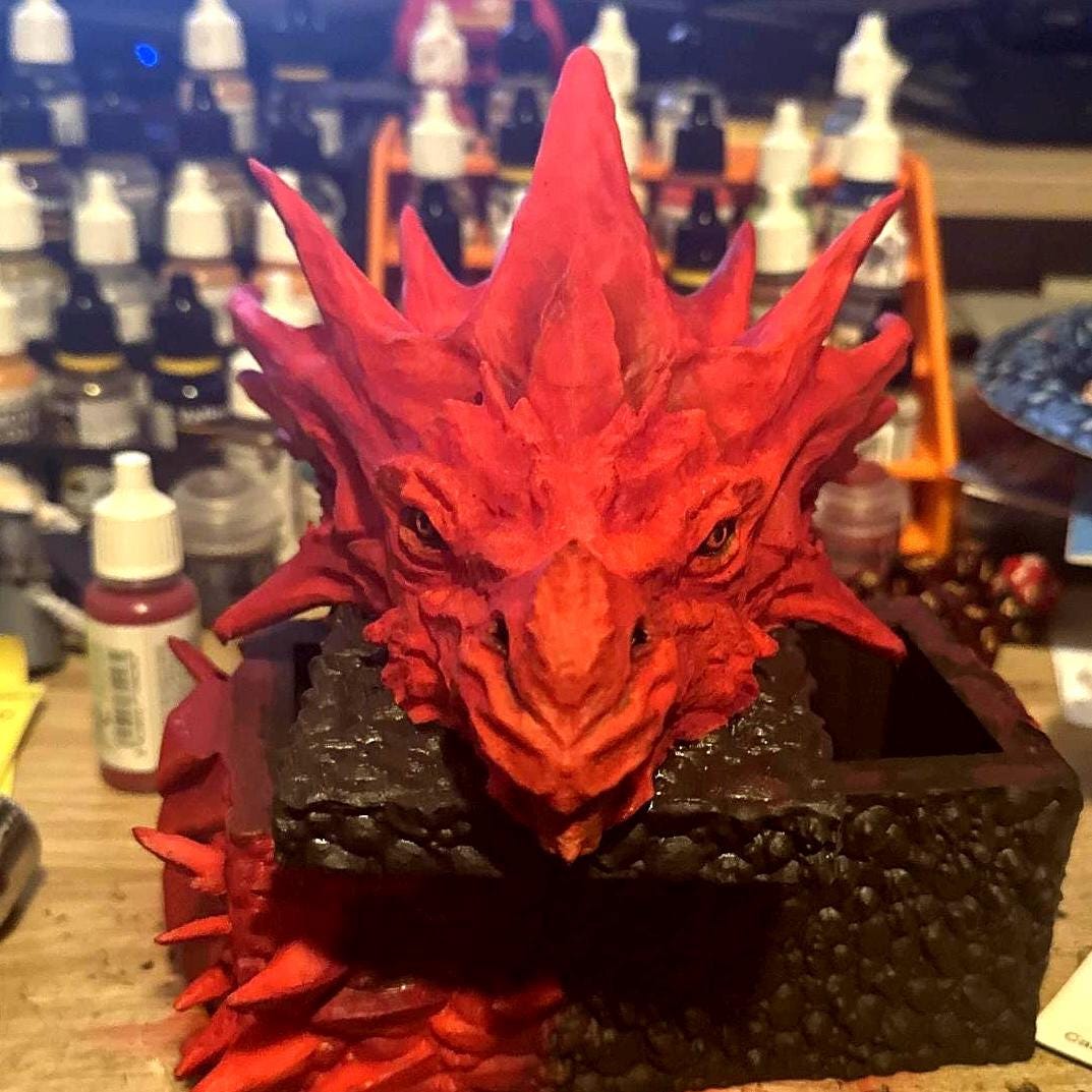 A red dragon head sculpture with a black base. The dragon has a textured surface and is painted in a bright red color. The dragon's eyes are black and the mouth is open slightly. The base is made of a dark, textured material. The sculpture is a decorative piece that would be perfect for any fantasy lover. Red dragon-themed dice case with resin dice set, perfect for DnD and RPG players. Stylish fantasy dice holder and storage box for tabletop gaming sessions.