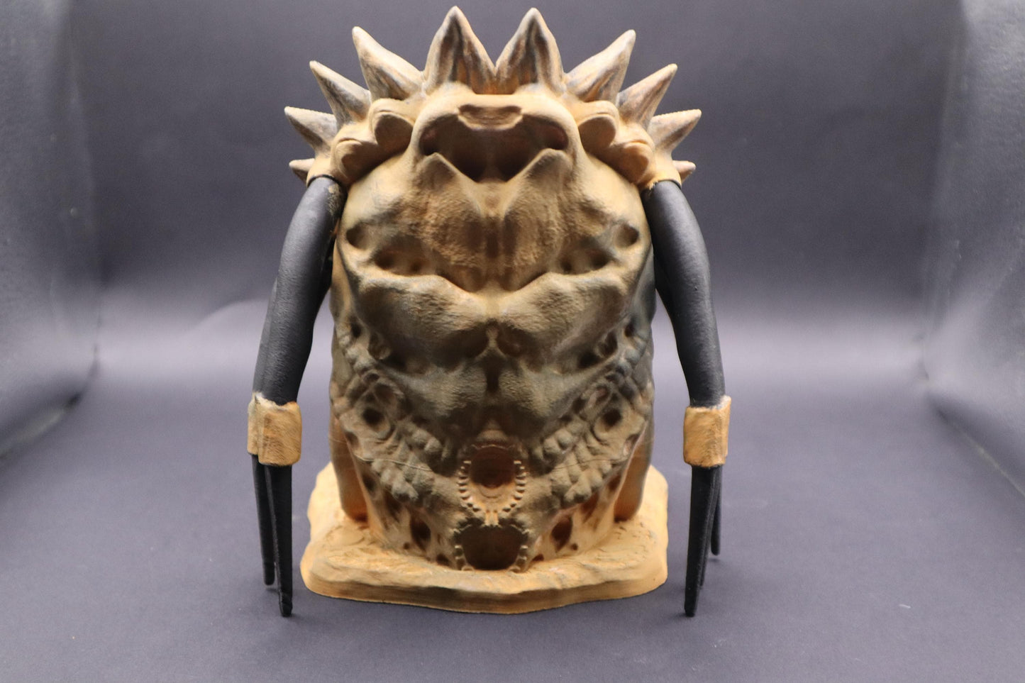 A Predator dice tower with a tan and brown color scheme. The tower features a detailed, textured design with a wide open mouth revealing pink gums and white teeth. The tower has black dreadlocks and a spiky head crest, showcasing the iconic alien creature inspired by the Predator movie franchise.