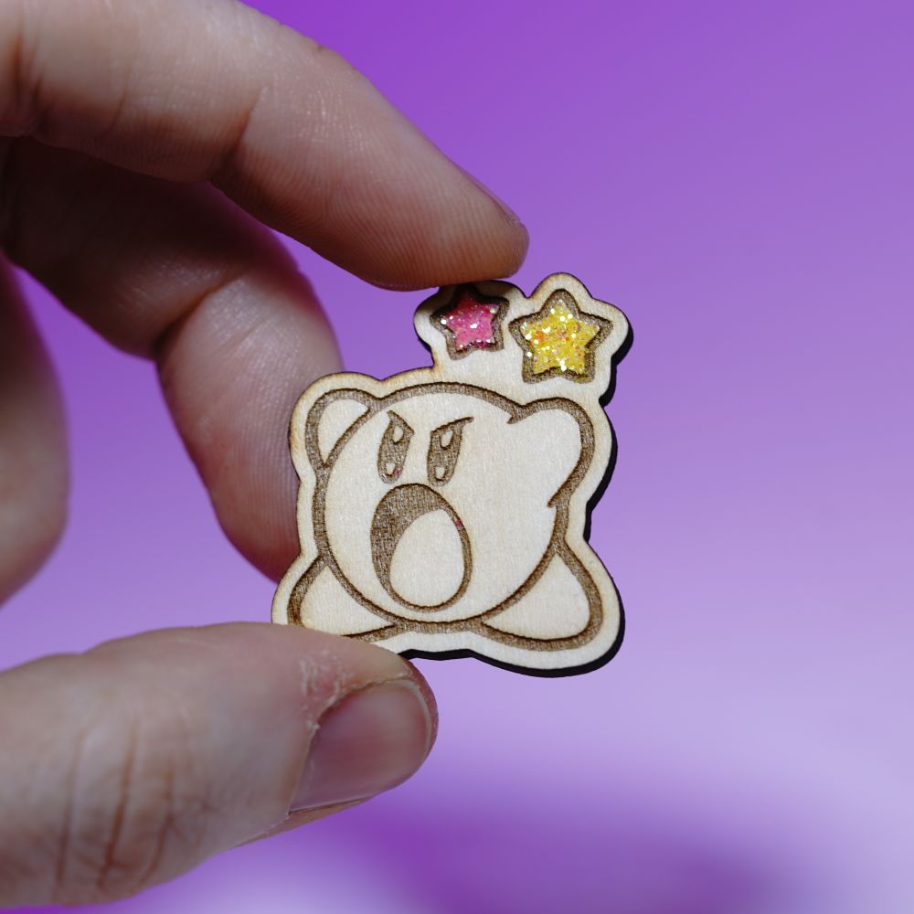 Cute Kirby wood pin with laser engraving and glittery star accents. Handmade gamer brooch for Nintendo fans, perfect for jackets or bags.