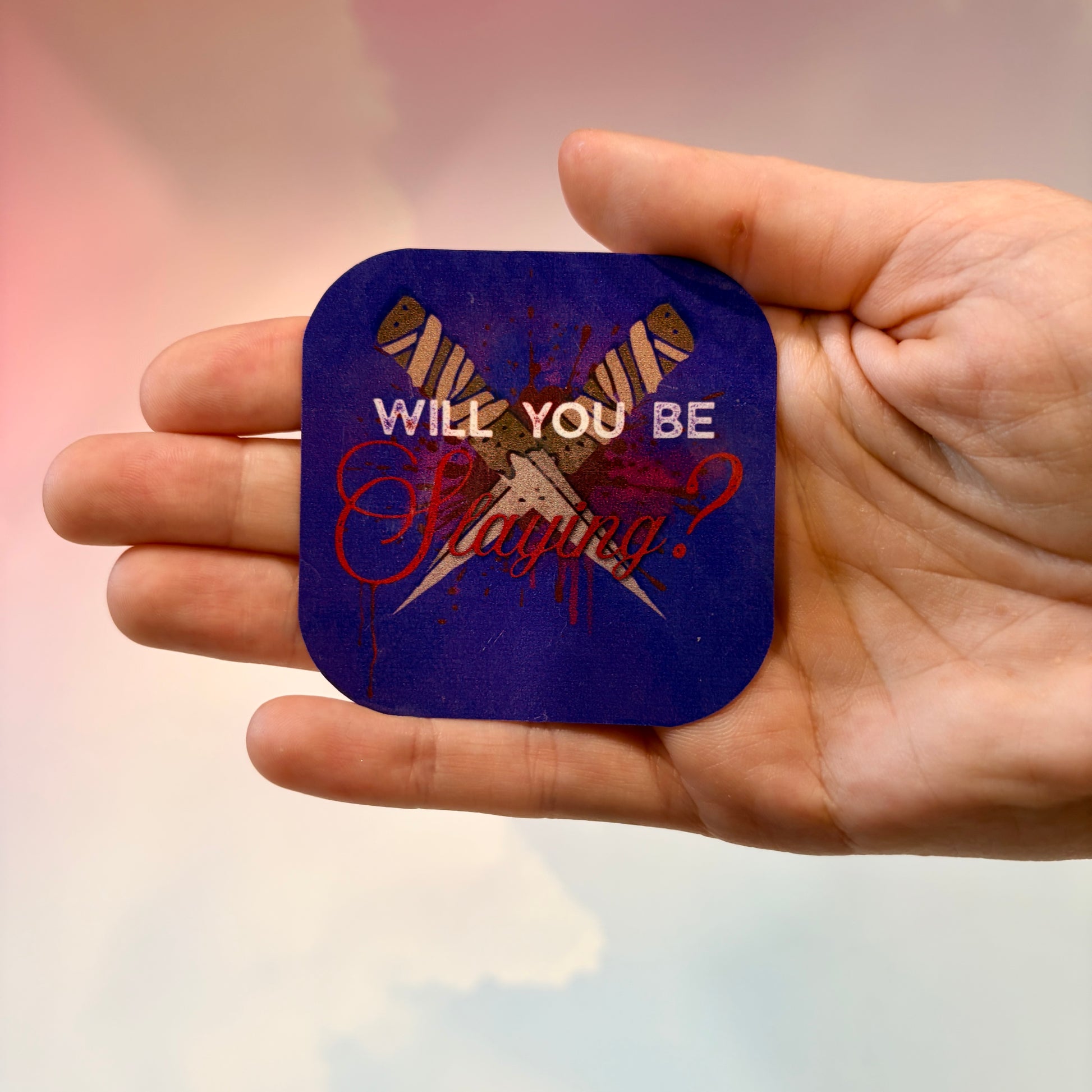 A hand holding a squarepurple sticker with rounded corners featuring a graphic of crossed stakes and the text 'WILL YOU BE Slaying?' in white and red.