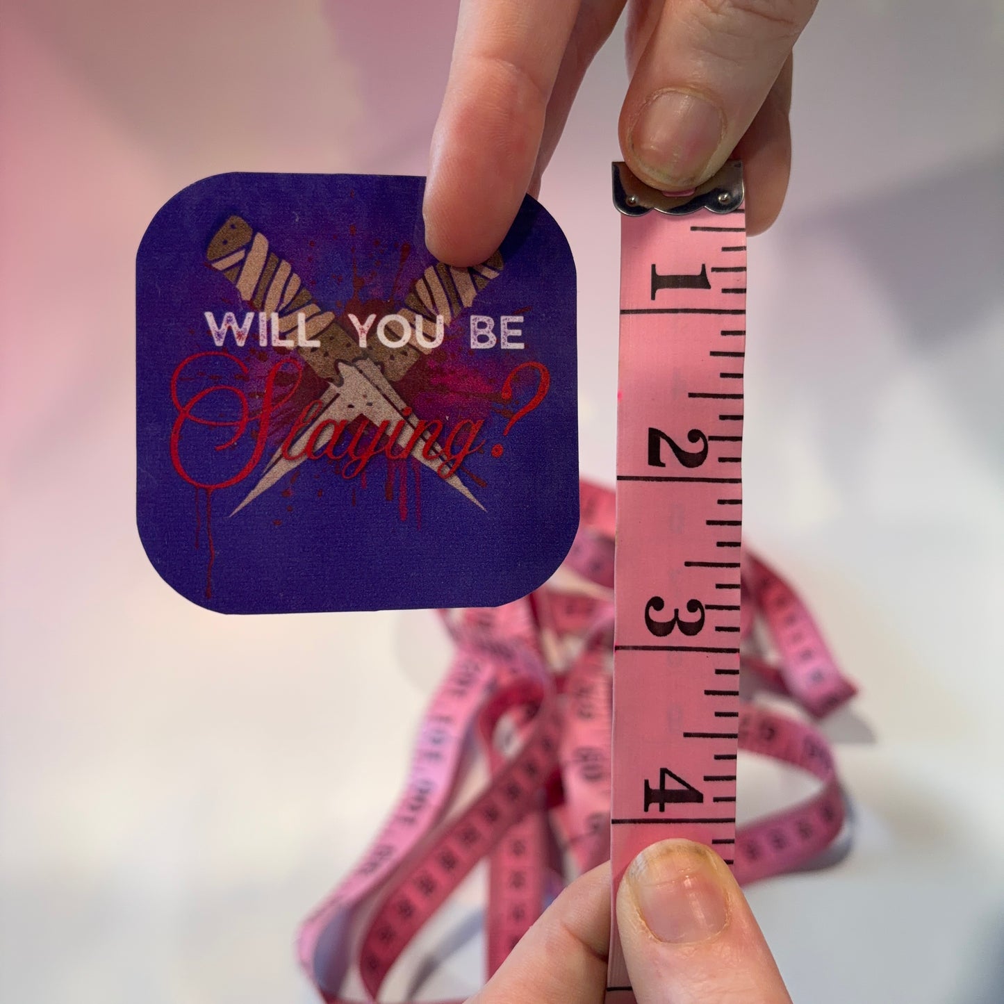A square purple sticker with rounded corners featuring a crossed stake design and the text 'WILL YOU BE Slaying?' in white and red script. A hand holds the sticker next to a pink measuring tape, with a pink measuring tape also visible in the blurred background.