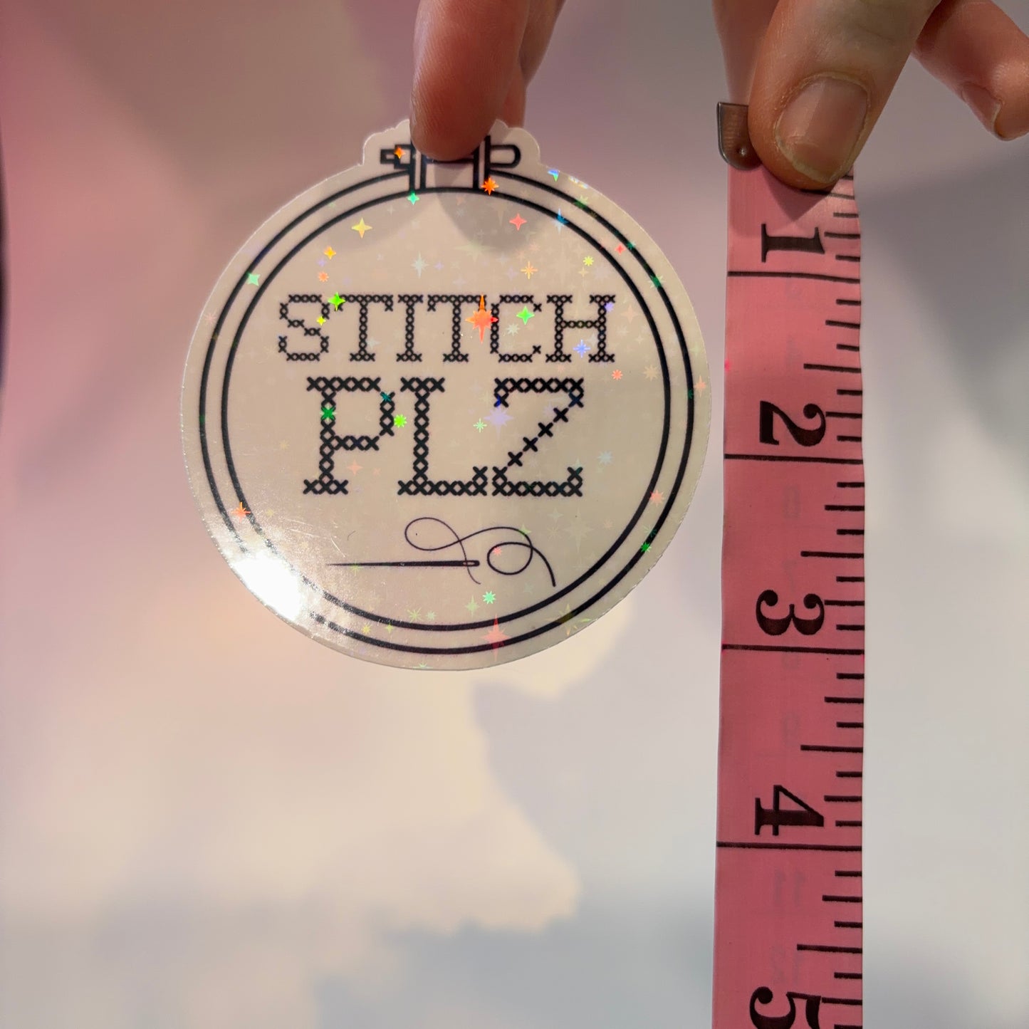 A round white sticker held by a hand, printed with the words “STITCH PLZ” in black stitched-style lettering inside a double-ring border. A pink measuring tape is shown next to it for scale.