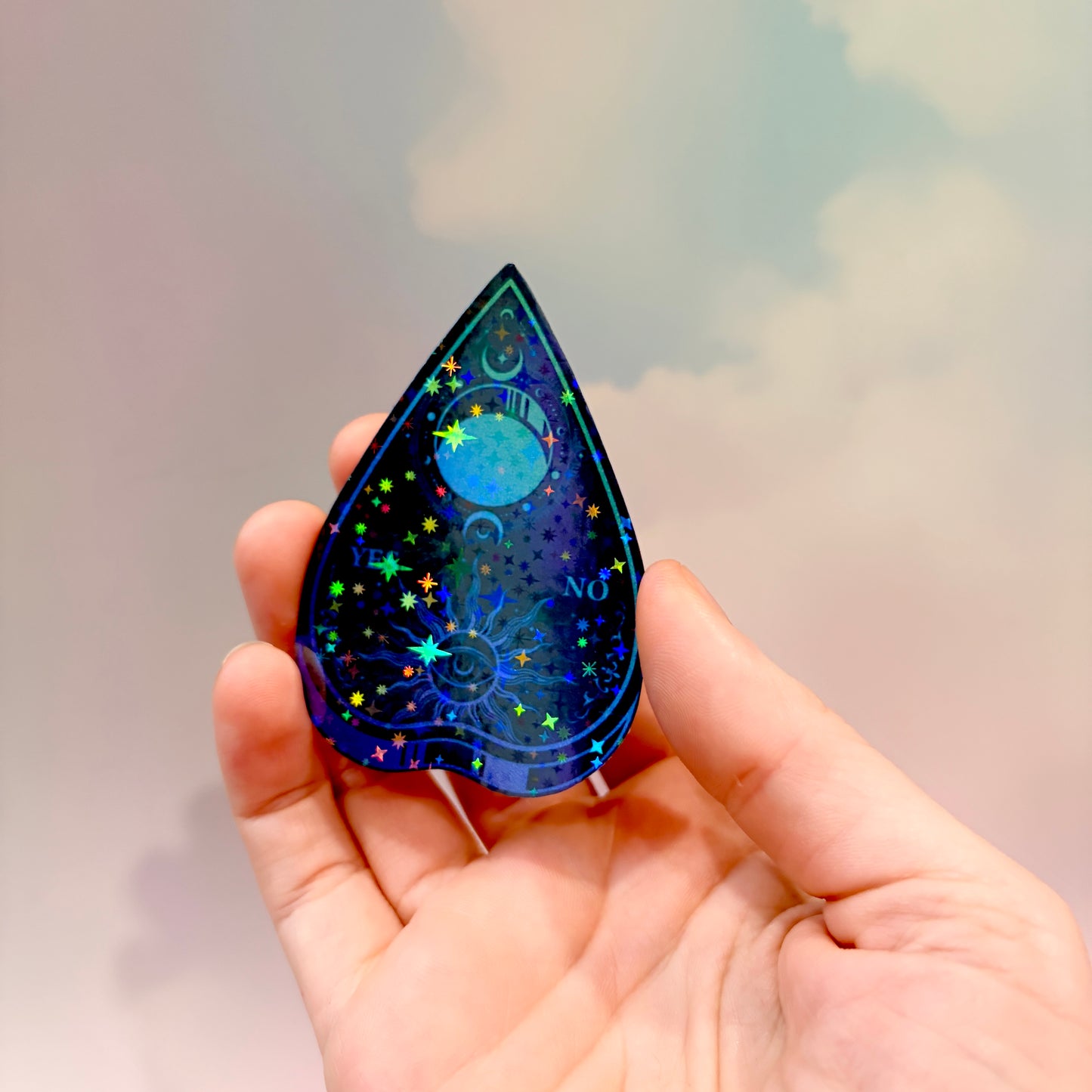 Black holographic Ouija planchette sticker with celestial sun, moon, stars, and blue-purple gradient artwork on water-resistant vinyl.