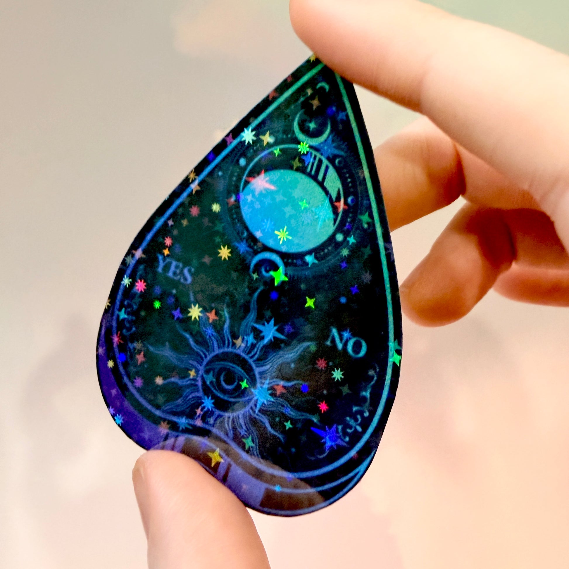Black holographic Ouija planchette sticker with celestial sun, moon, stars, and blue-purple gradient artwork on water-resistant vinyl.
