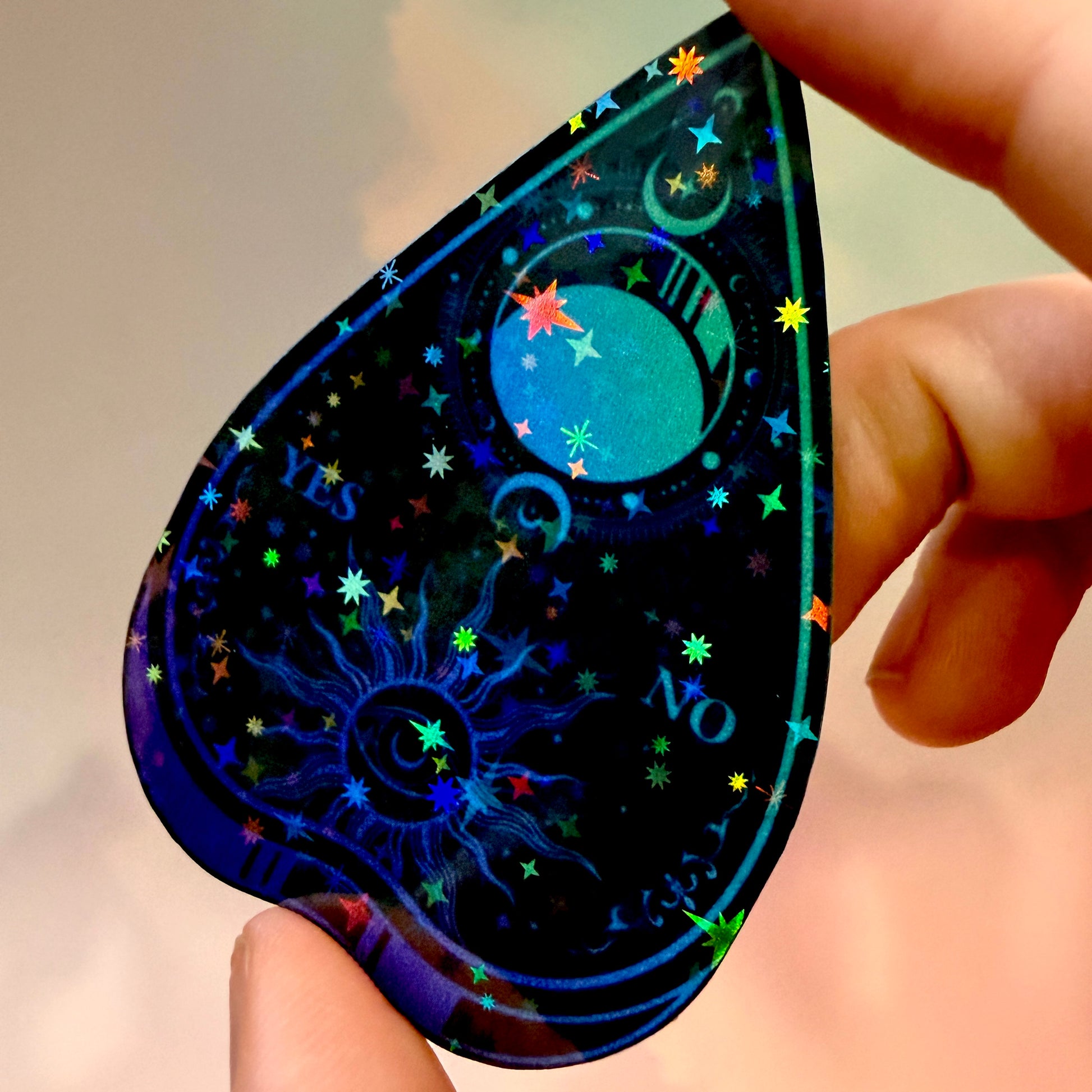 Black holographic Ouija planchette sticker with celestial sun, moon, stars, and blue-purple gradient artwork on water-resistant vinyl.
