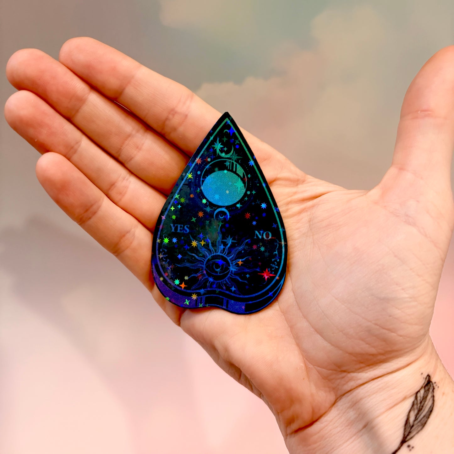 Black holographic Ouija planchette sticker with celestial sun, moon, stars, and blue-purple gradient artwork on water-resistant vinyl.