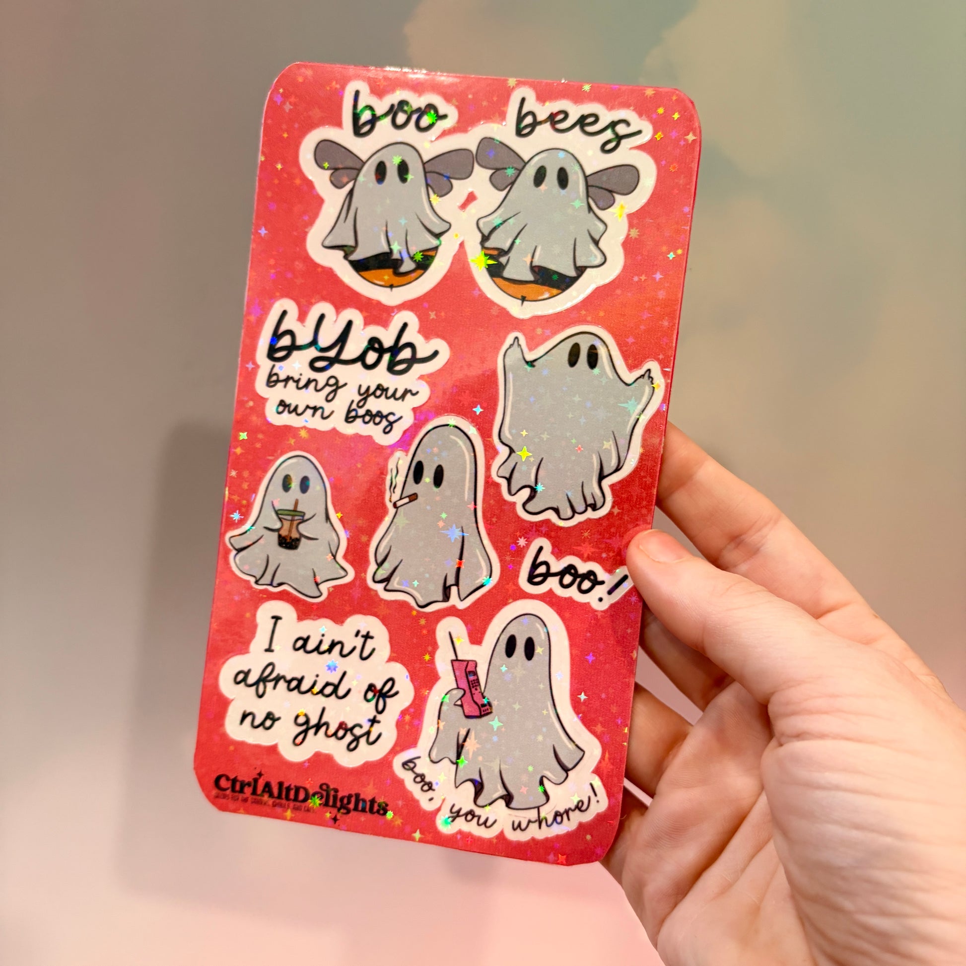 Holographic sticker sheet featuring eight cute ghost vinyl stickers with funny sayings including "Boo Bees," "BYOB Bring Your Own Boos," "I Ain't Afraid of No Ghost," and other snarky spooky designs on a pink glitter background.