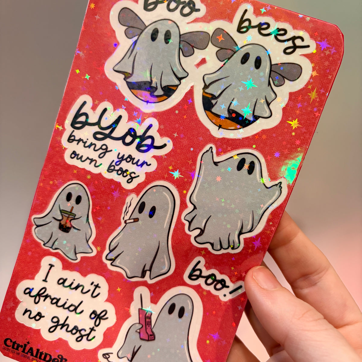 Holographic sticker sheet featuring eight cute ghost vinyl stickers with funny sayings including "Boo Bees," "BYOB Bring Your Own Boos," "I Ain't Afraid of No Ghost," and other snarky spooky designs on a pink glitter background.