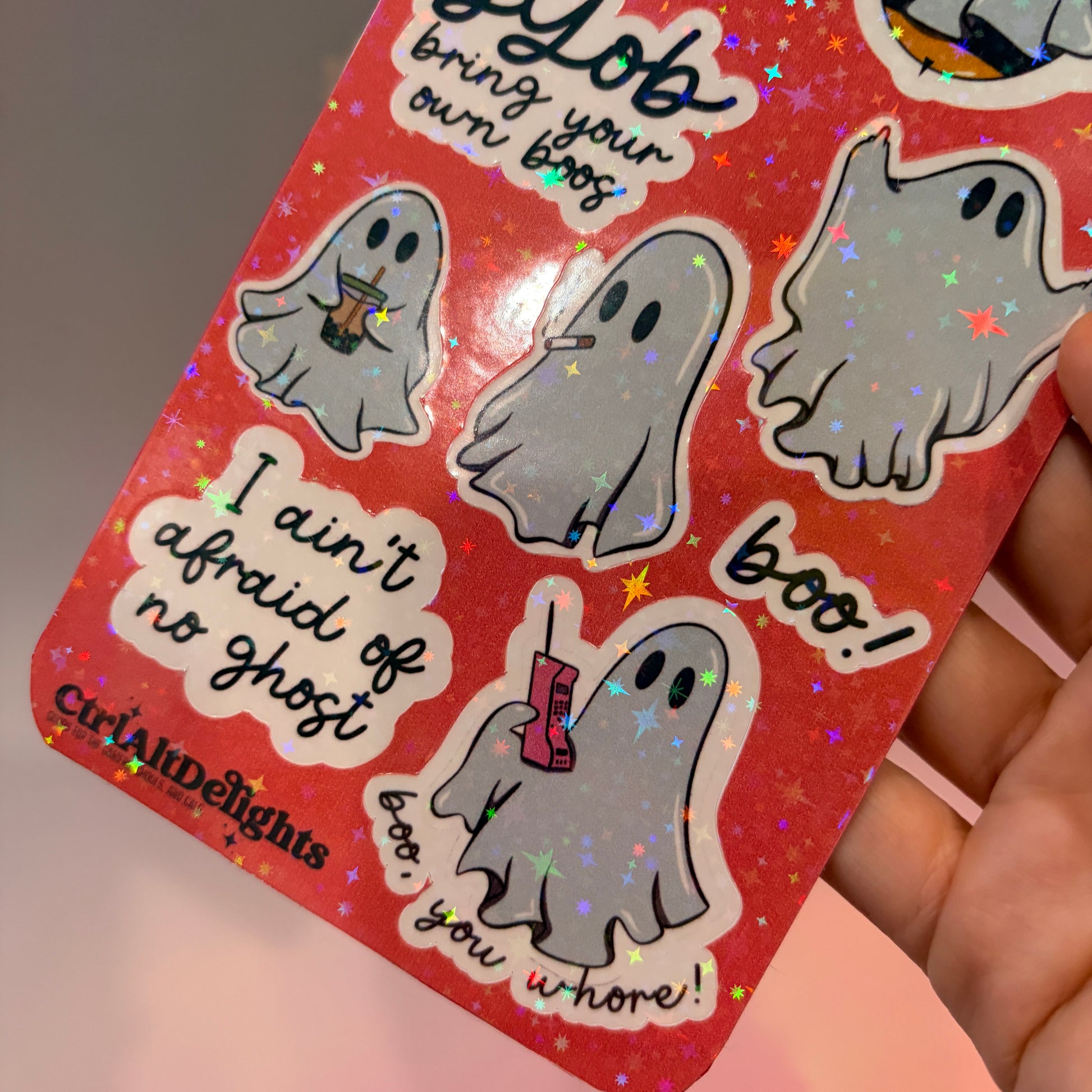 Holographic sticker sheet featuring eight cute ghost vinyl stickers with funny sayings including "Boo Bees," "BYOB Bring Your Own Boos," "I Ain't Afraid of No Ghost," and other snarky spooky designs on a pink glitter background.