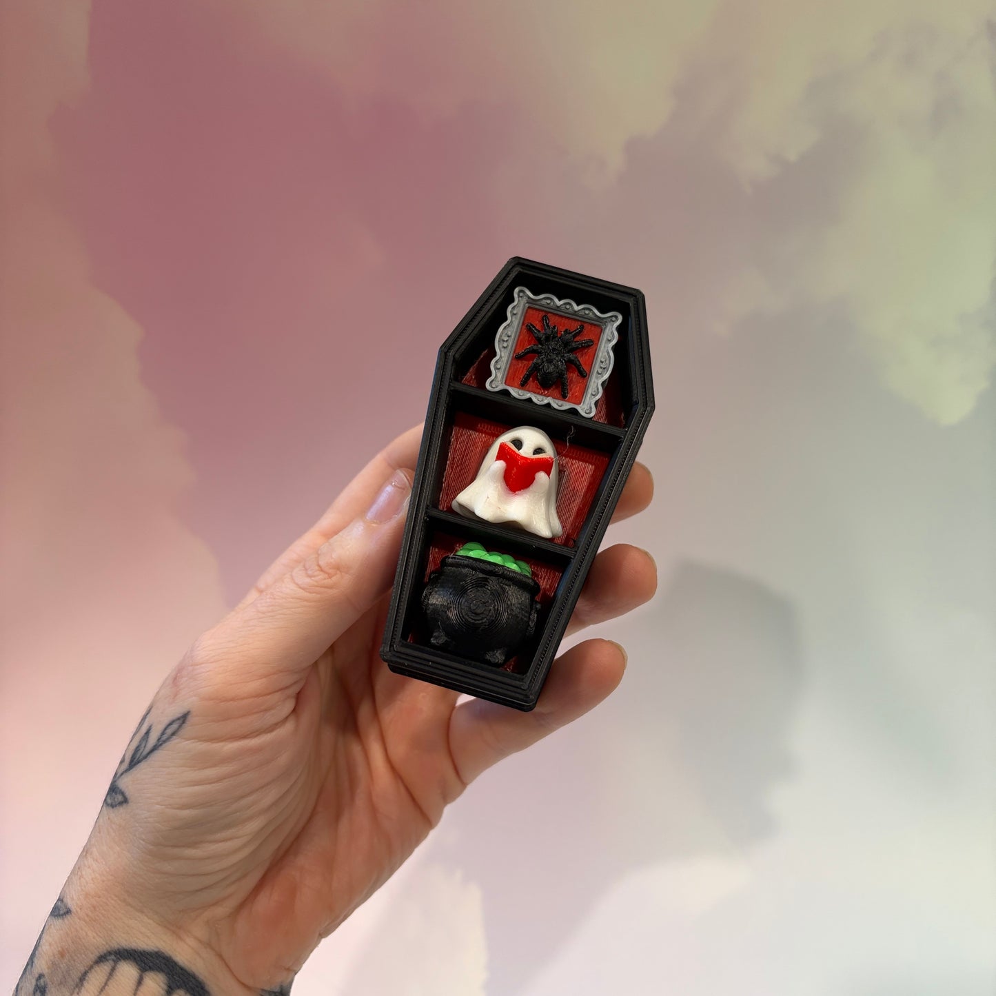 Mini 3D printed black coffin shelf fidget clicker with silver gothic trim and red interior. Features three decorative keycaps: a framed black spider, a ghost reading a book, and a bubbling green cauldron. Includes a silver keychain and is available with clicky or silent switches.