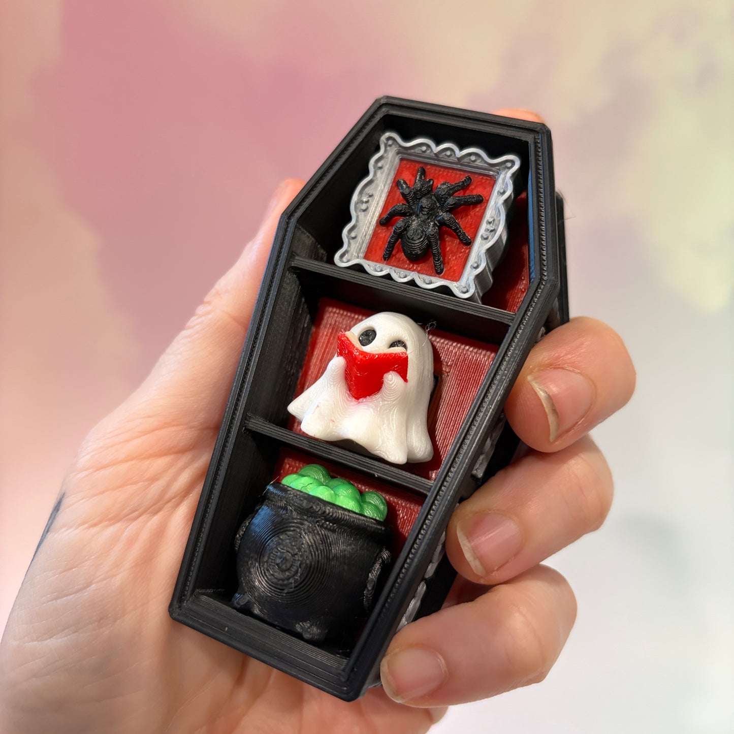 Mini 3D printed black coffin shelf fidget clicker with silver gothic trim and red interior. Features three decorative keycaps: a framed black spider, a ghost reading a book, and a bubbling green cauldron. Includes a silver keychain and is available with clicky or silent switches.