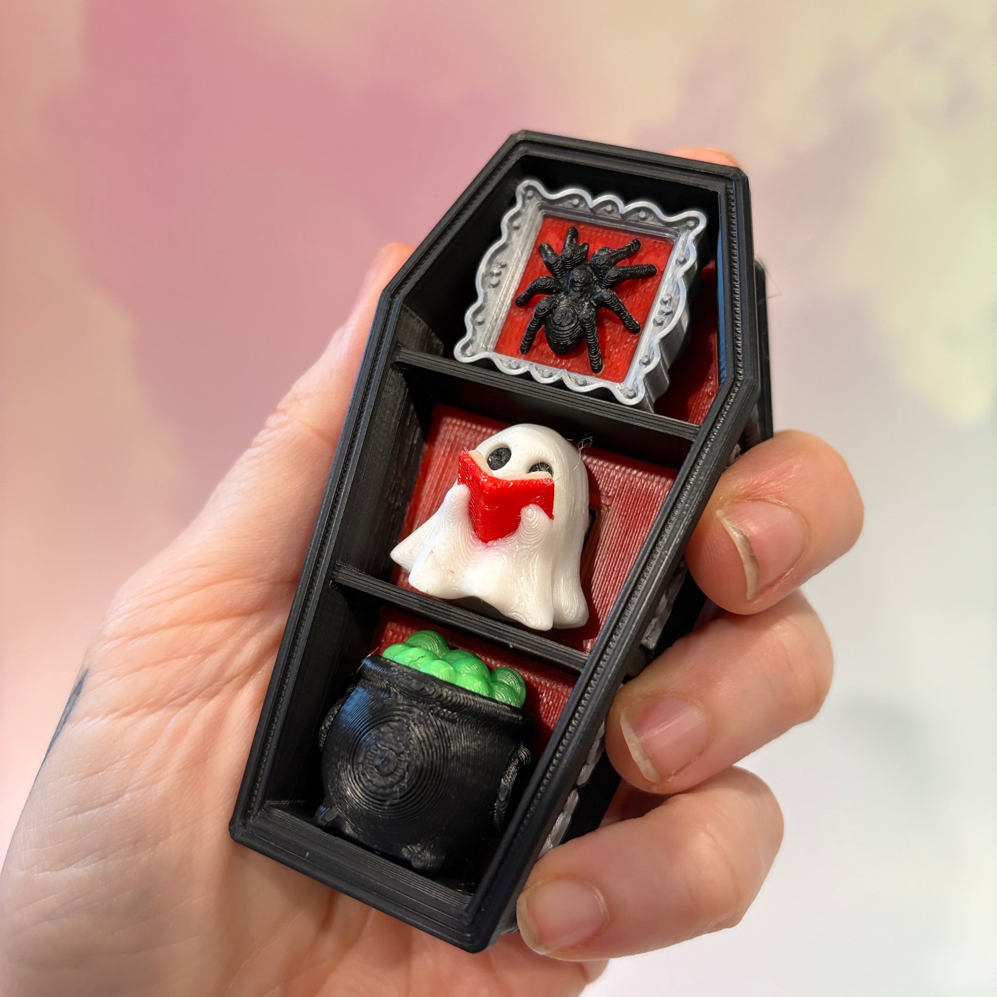Mini 3D printed black coffin shelf fidget clicker with silver gothic trim and red interior. Features three decorative keycaps: a framed black spider, a ghost reading a book, and a bubbling green cauldron. Includes a silver keychain and is available with clicky or silent switches.