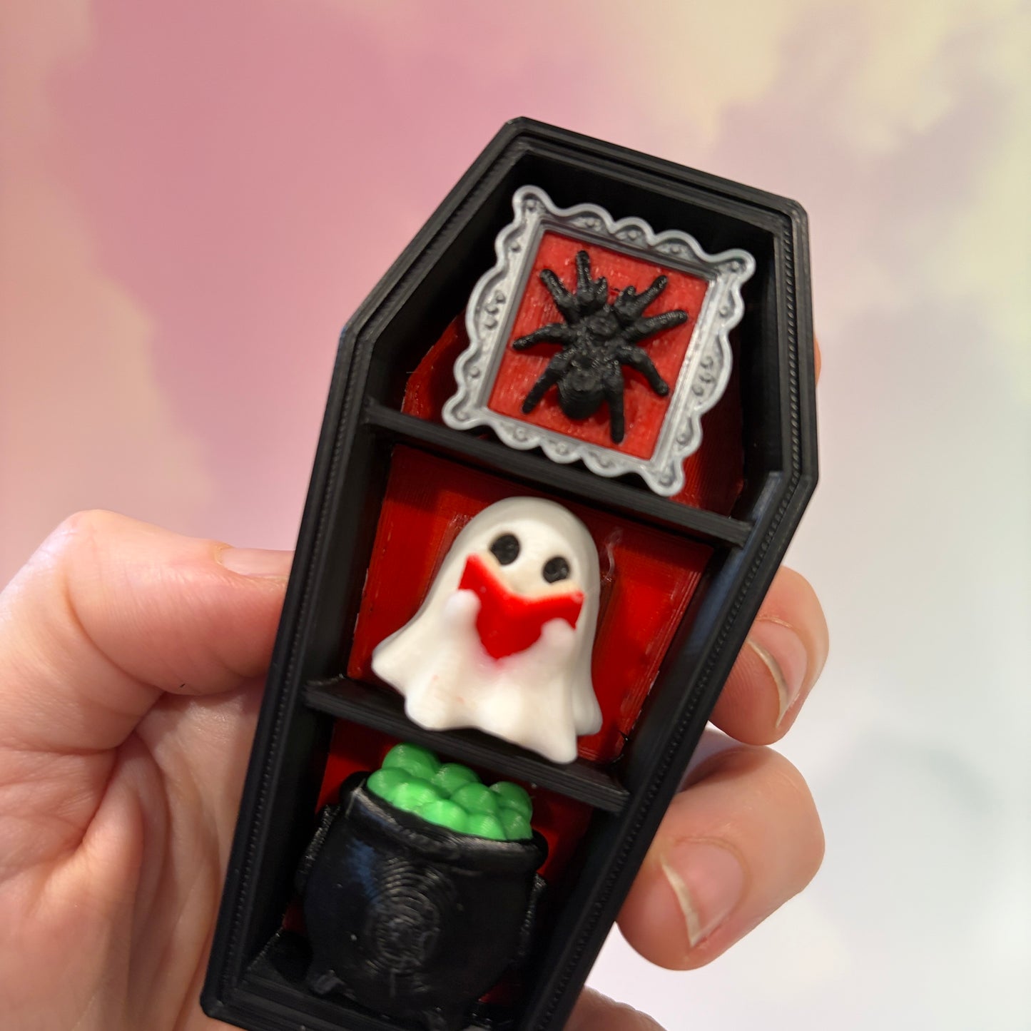 Mini 3D printed black coffin shelf fidget clicker with silver gothic trim and red interior. Features three decorative keycaps: a framed black spider, a ghost reading a book, and a bubbling green cauldron. Includes a silver keychain and is available with clicky or silent switches.