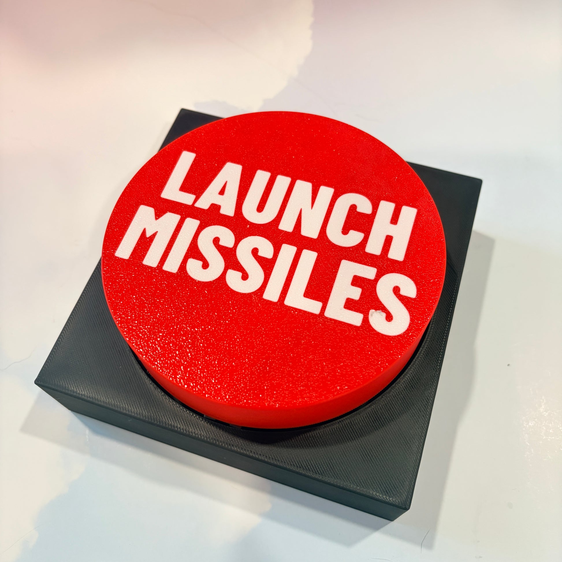 Oversized red 3D printed fidget button with white “LAUNCH MISSILES” lettering on a black square base, shown on a white surface.
