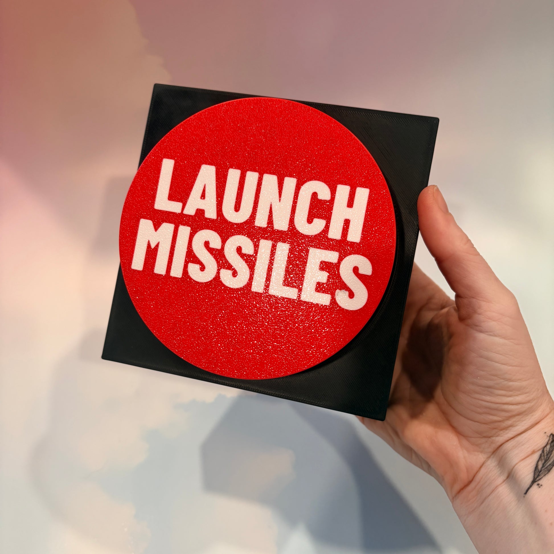 Oversized red 3D printed fidget button with white “LAUNCH MISSILES” lettering on a black square base, shown on a white surface.