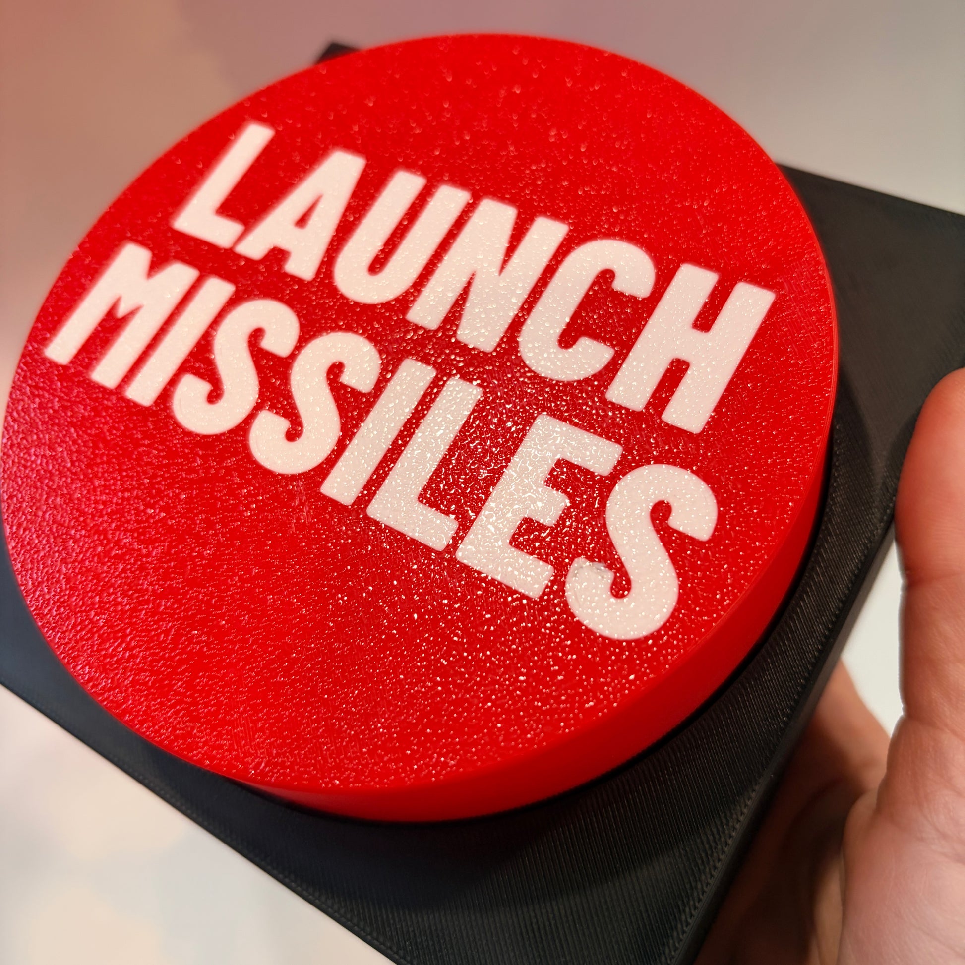 Oversized red 3D printed fidget button with white “LAUNCH MISSILES” lettering on a black square base, shown on a white surface.
