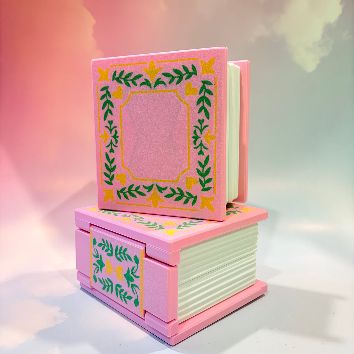 A pink decorative book-shaped box with a hinged cover, shown open and closed in a stacked arrangement. The cover and front panel feature a yellow border with green leafy vine motifs, and the pages are off-white.