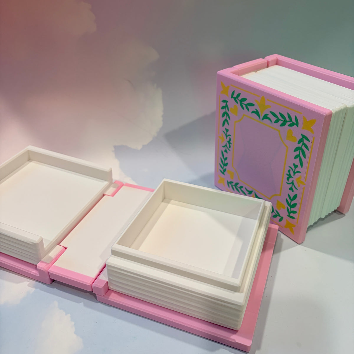 A set of miniature book-shaped storage boxes or trays in pastel pink and cream, with one standing upright on the right decorated with a green and yellow floral border and one lying open on the left.