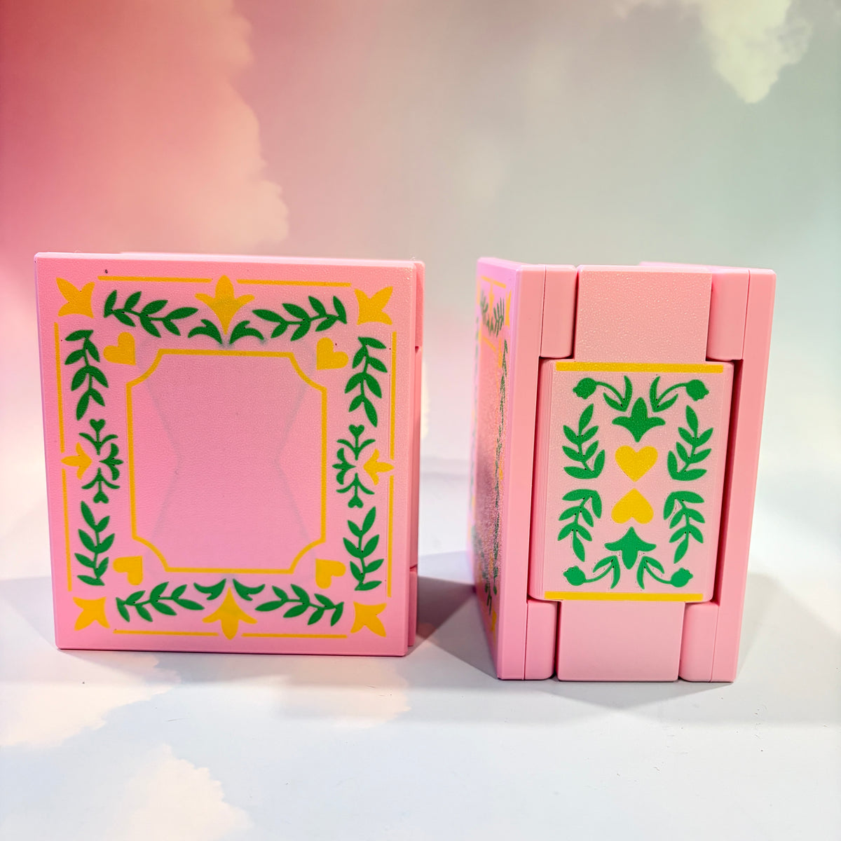 A pink plastic toy box with decorative green and yellow leaf and heart patterns, shown in two views against a pastel pink and white background.