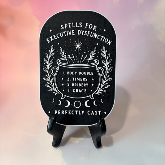 A 3D printed witchy sign titled “Spells for Executive Dysfunction,” featuring a black rounded rectangular base with raised white lettering and intricate white magical artwork. At the top, the phrase “Spells for Executive Dysfunction” curves around small decorative stars. In the center, a large outlined witch’s cauldron holds a list of four helpful spells.