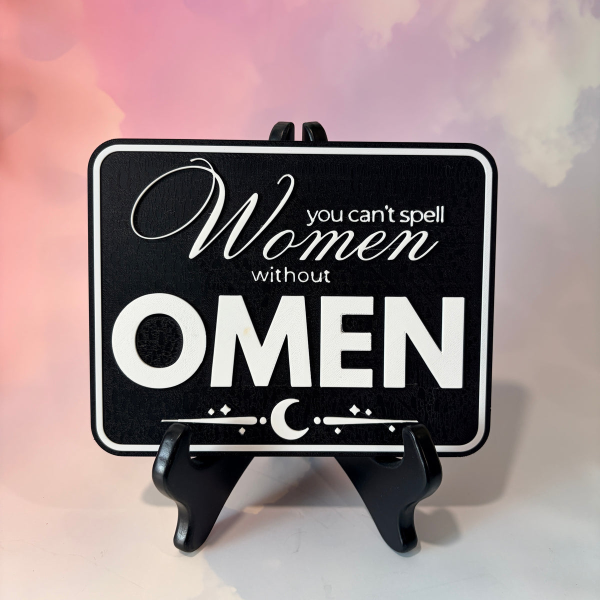 A rectangular 3D printed decorative sign featuring the phrase “You Can’t Spell Women Without Omen” in raised white lettering against a black background.
