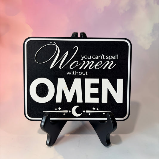 A rectangular 3D printed decorative sign featuring the phrase “You Can’t Spell Women Without Omen” in raised white lettering against a black background.