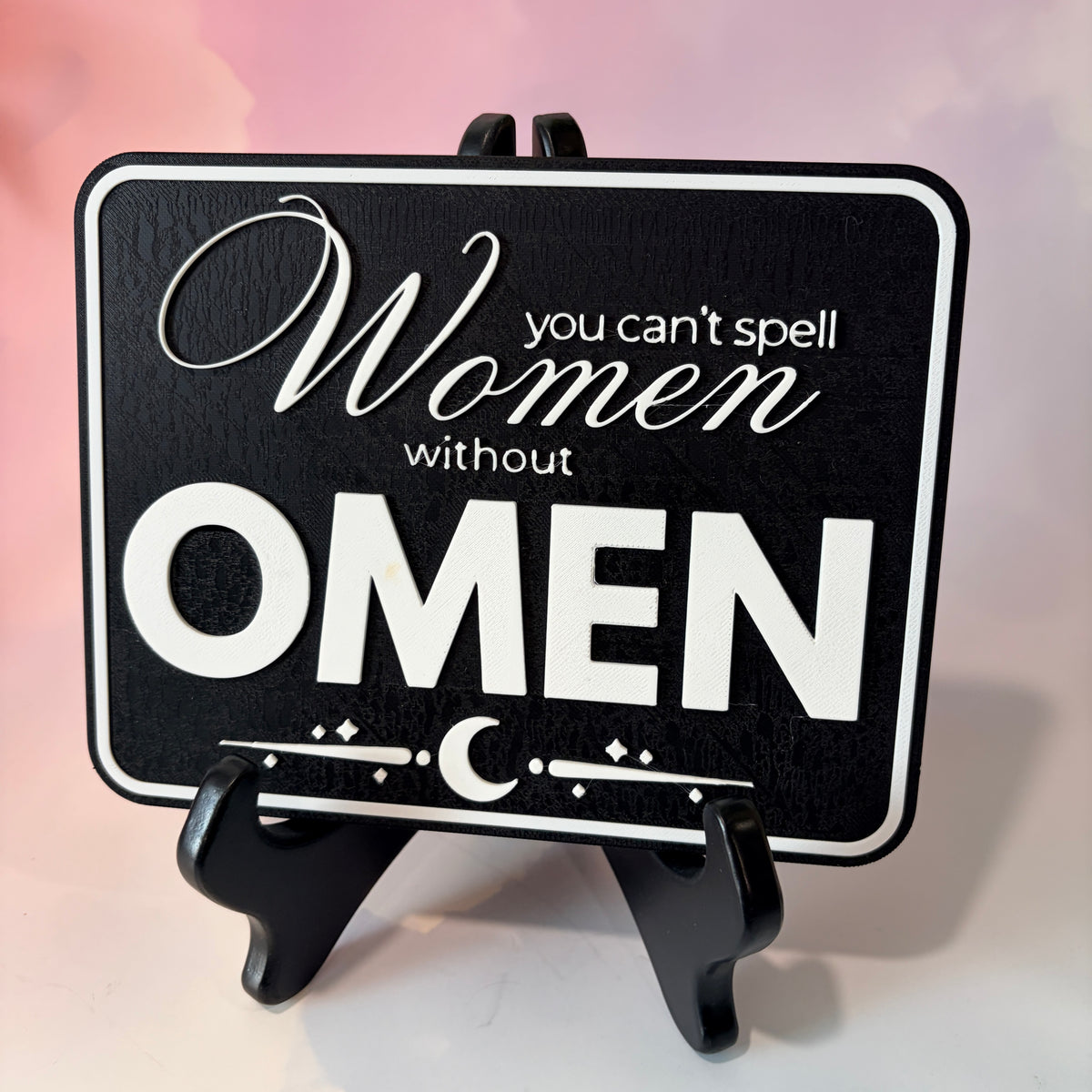 A rectangular 3D printed decorative sign featuring the phrase “You Can’t Spell Women Without Omen” in raised white lettering against a black background.