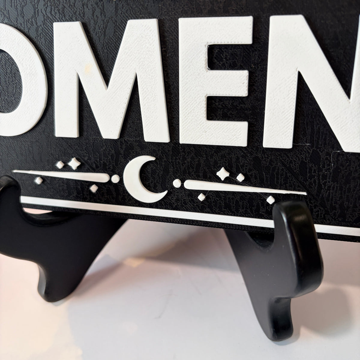 A rectangular 3D printed decorative sign featuring the phrase “You Can’t Spell Women Without Omen” in raised white lettering against a black background.