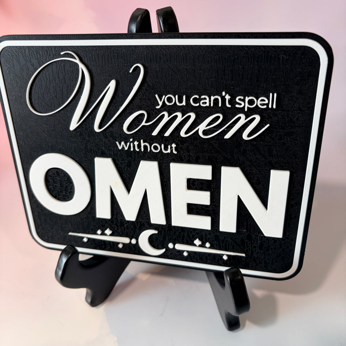 A rectangular 3D printed decorative sign featuring the phrase “You Can’t Spell Women Without Omen” in raised white lettering against a black background.
