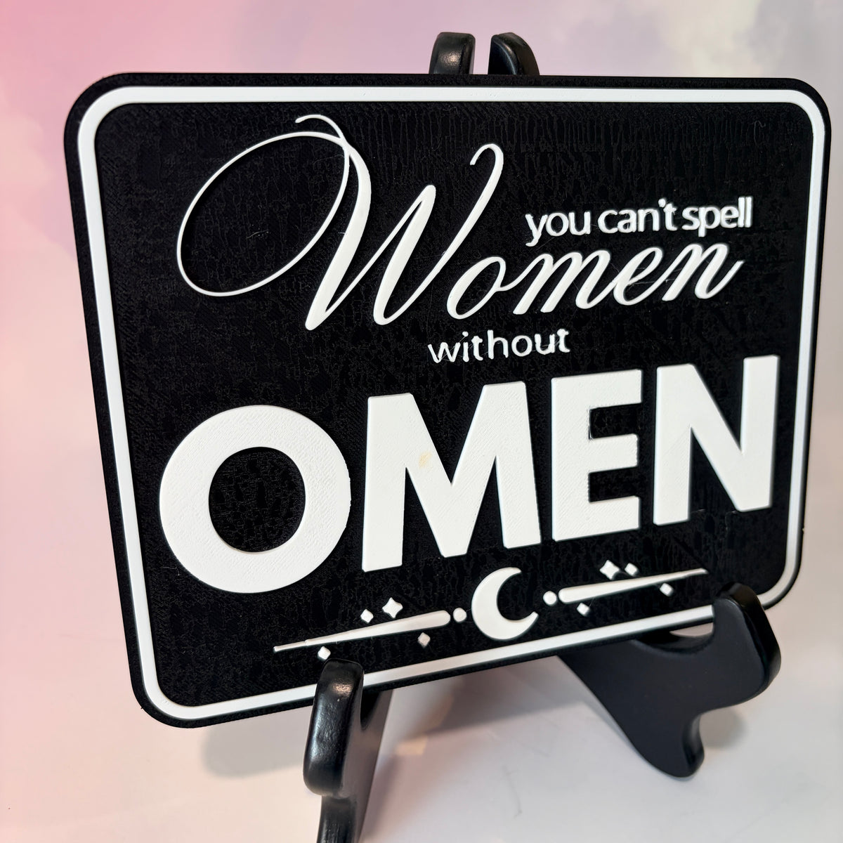A rectangular 3D printed decorative sign featuring the phrase “You Can’t Spell Women Without Omen” in raised white lettering against a black background.