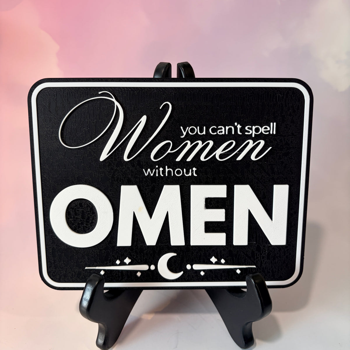 A rectangular 3D printed decorative sign featuring the phrase “You Can’t Spell Women Without Omen” in raised white lettering against a black background.