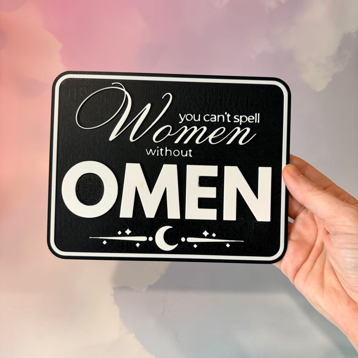 A rectangular 3D printed decorative sign featuring the phrase “You Can’t Spell Women Without Omen” in raised white lettering against a black background.