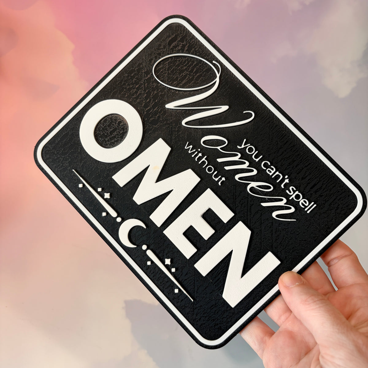 A rectangular 3D printed decorative sign featuring the phrase “You Can’t Spell Women Without Omen” in raised white lettering against a black background.