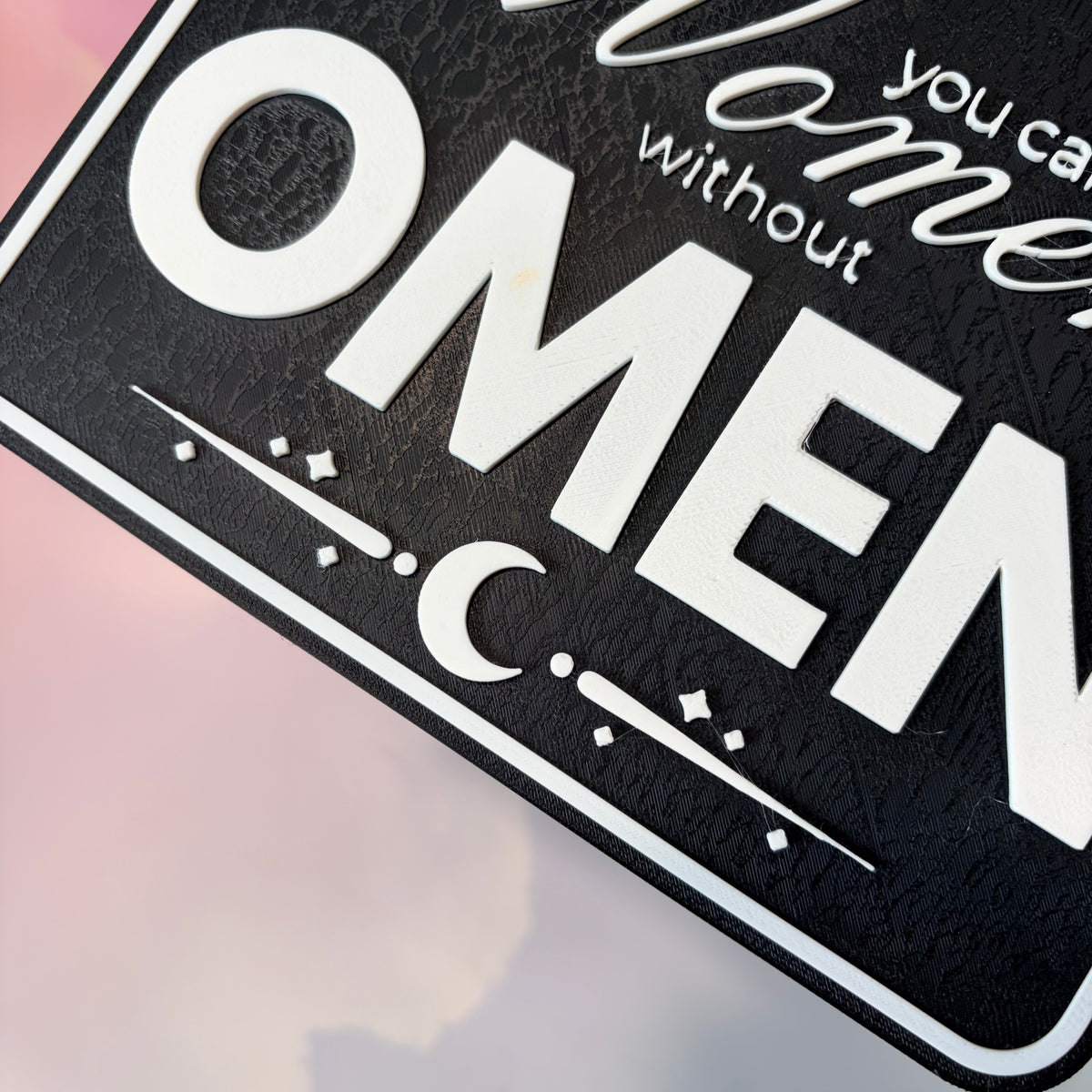 A rectangular 3D printed decorative sign featuring the phrase “You Can’t Spell Women Without Omen” in raised white lettering against a black background.