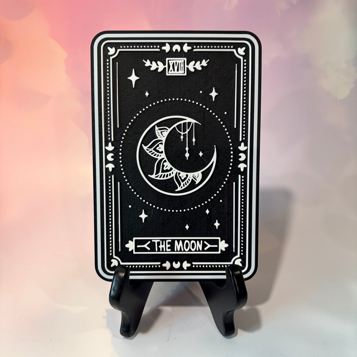 A vertical 3D printed tarot card sign featuring The Moon from the Major Arcana, displayed upright on a small black easel against a soft pink, peach, and lavender background. All 22 Major Arcana tarot card designs are available, allowing customers to choose a card that reflects their interests, spiritual practice, or personal journey.