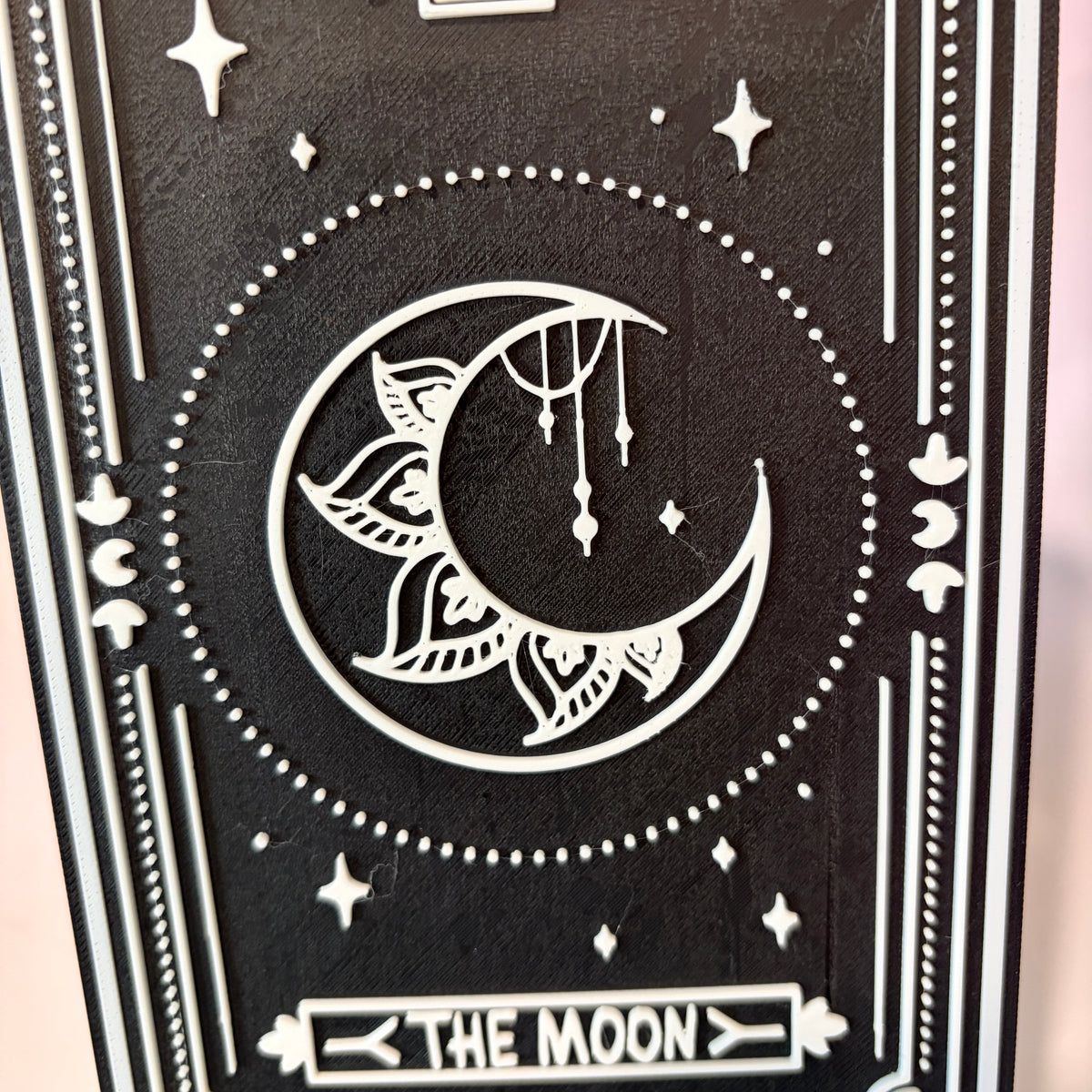 A vertical 3D printed tarot card sign featuring The Moon from the Major Arcana, displayed upright on a small black easel against a soft pink, peach, and lavender background. All 22 Major Arcana tarot card designs are available, allowing customers to choose a card that reflects their interests, spiritual practice, or personal journey.