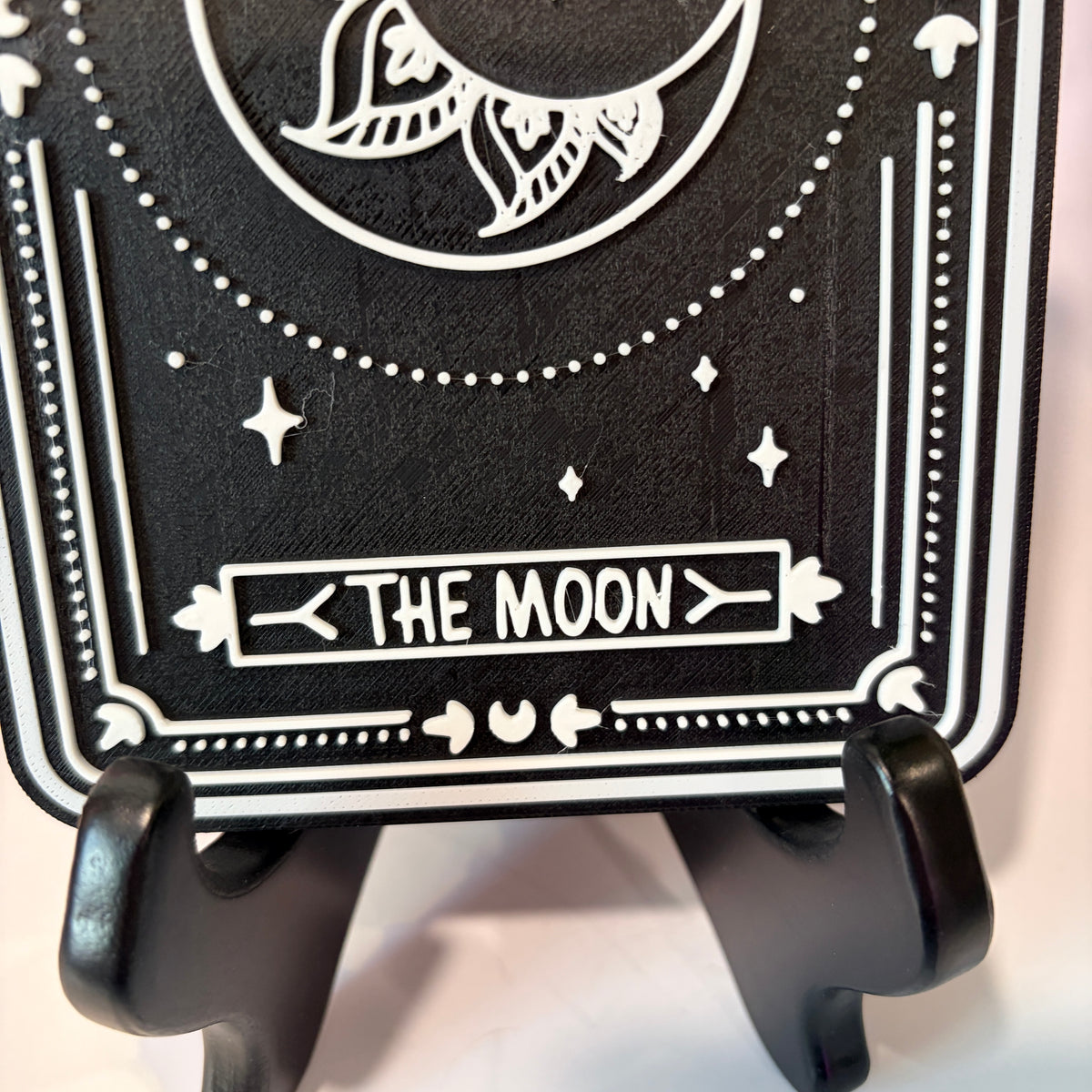 A vertical 3D printed tarot card sign featuring The Moon from the Major Arcana, displayed upright on a small black easel against a soft pink, peach, and lavender background. All 22 Major Arcana tarot card designs are available, allowing customers to choose a card that reflects their interests, spiritual practice, or personal journey.
