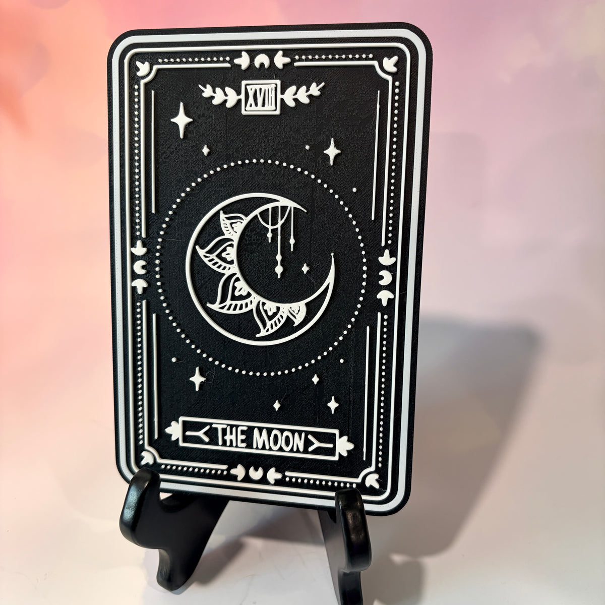 A vertical 3D printed tarot card sign featuring The Moon from the Major Arcana, displayed upright on a small black easel against a soft pink, peach, and lavender background. All 22 Major Arcana tarot card designs are available, allowing customers to choose a card that reflects their interests, spiritual practice, or personal journey.