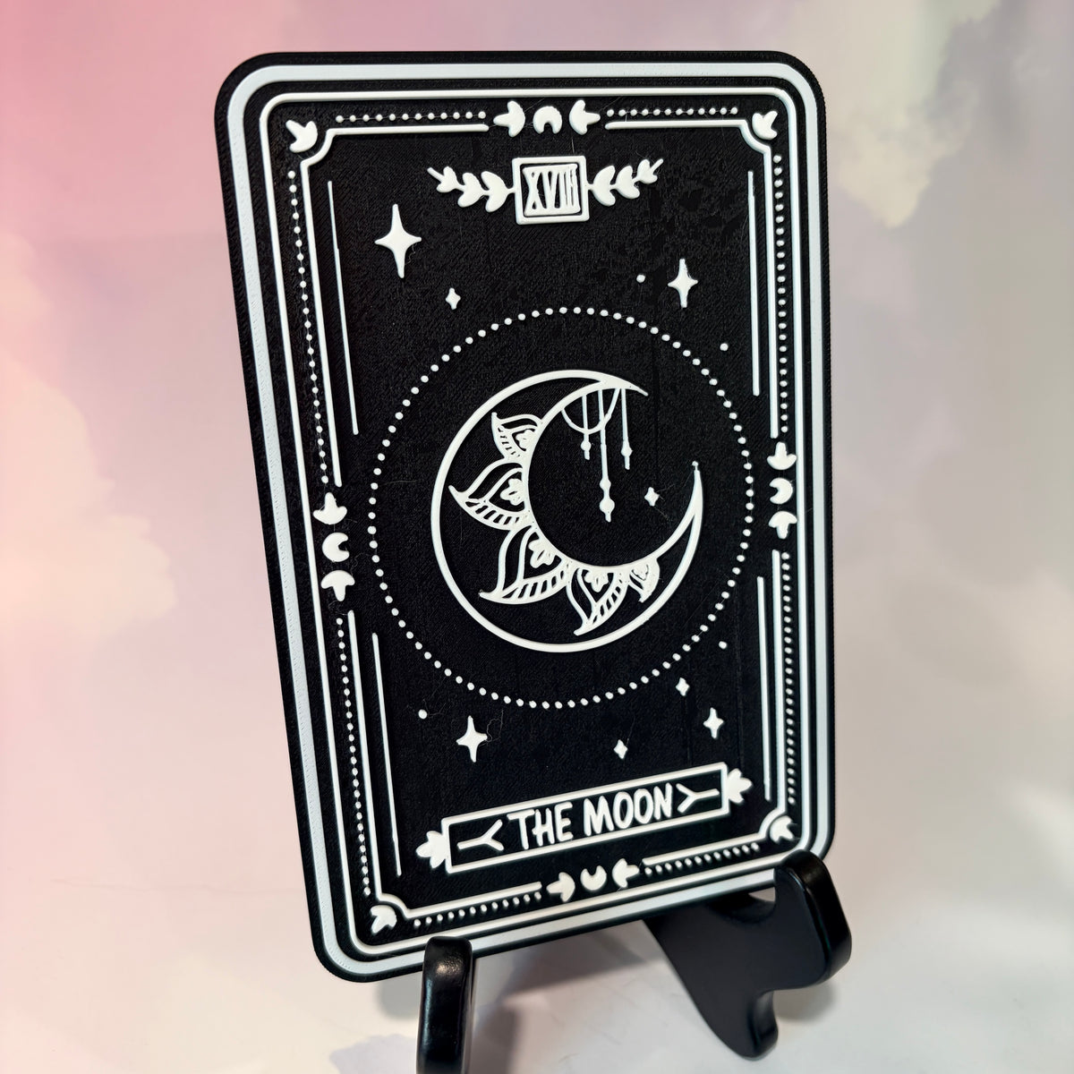 A vertical 3D printed tarot card sign featuring The Moon from the Major Arcana, displayed upright on a small black easel against a soft pink, peach, and lavender background. All 22 Major Arcana tarot card designs are available, allowing customers to choose a card that reflects their interests, spiritual practice, or personal journey.