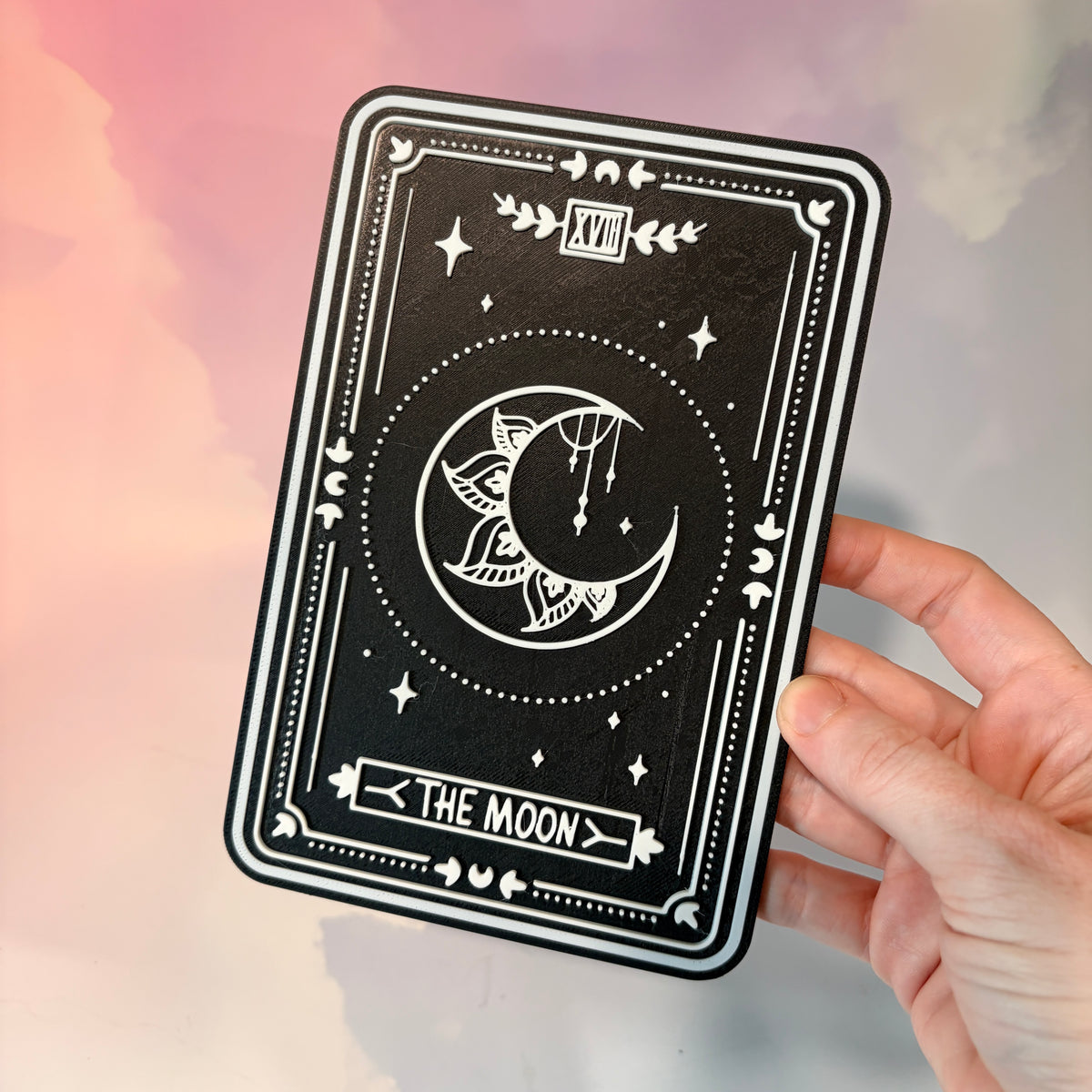 A vertical 3D printed tarot card sign featuring The Moon from the Major Arcana, displayed upright on a small black easel against a soft pink, peach, and lavender background. All 22 Major Arcana tarot card designs are available, allowing customers to choose a card that reflects their interests, spiritual practice, or personal journey.