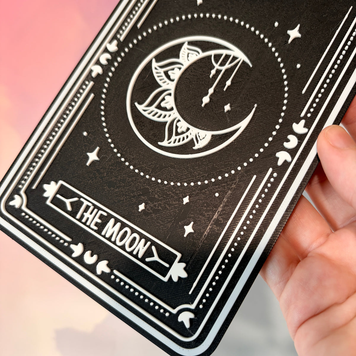 A vertical 3D printed tarot card sign featuring The Moon from the Major Arcana, displayed upright on a small black easel against a soft pink, peach, and lavender background. All 22 Major Arcana tarot card designs are available, allowing customers to choose a card that reflects their interests, spiritual practice, or personal journey.