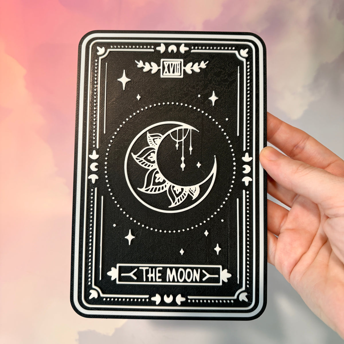 A vertical 3D printed tarot card sign featuring The Moon from the Major Arcana, displayed upright on a small black easel against a soft pink, peach, and lavender background. All 22 Major Arcana tarot card designs are available, allowing customers to choose a card that reflects their interests, spiritual practice, or personal journey.