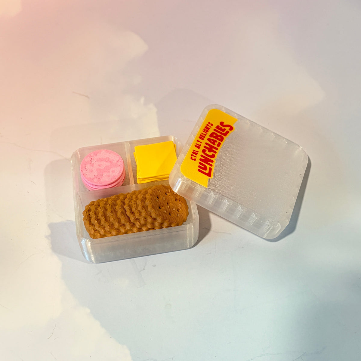 3D printed mini snack box fidget clicker with ham, cheese, and crackers under a semi-transparent lid. Ham and cheese have soft silent presses, while the crackers make satisfying audible clicks.