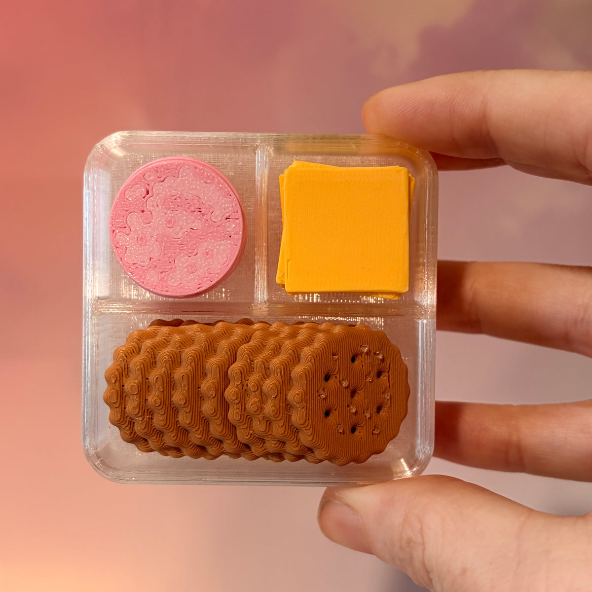 3D printed mini snack box fidget clicker with ham, cheese, and crackers under a semi-transparent lid. Ham and cheese have soft silent presses, while the crackers make satisfying audible clicks.