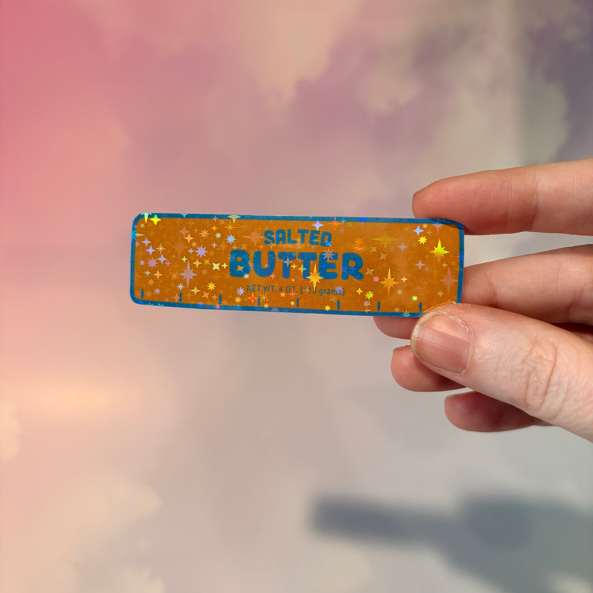 Holographic vinyl sticker shaped like a classic stick of butter, measuring approximately 3 inches long by 1 inch tall. Colorful reflective finish adds a playful touch to laptops, notebooks, tumblers, and sticker collections.