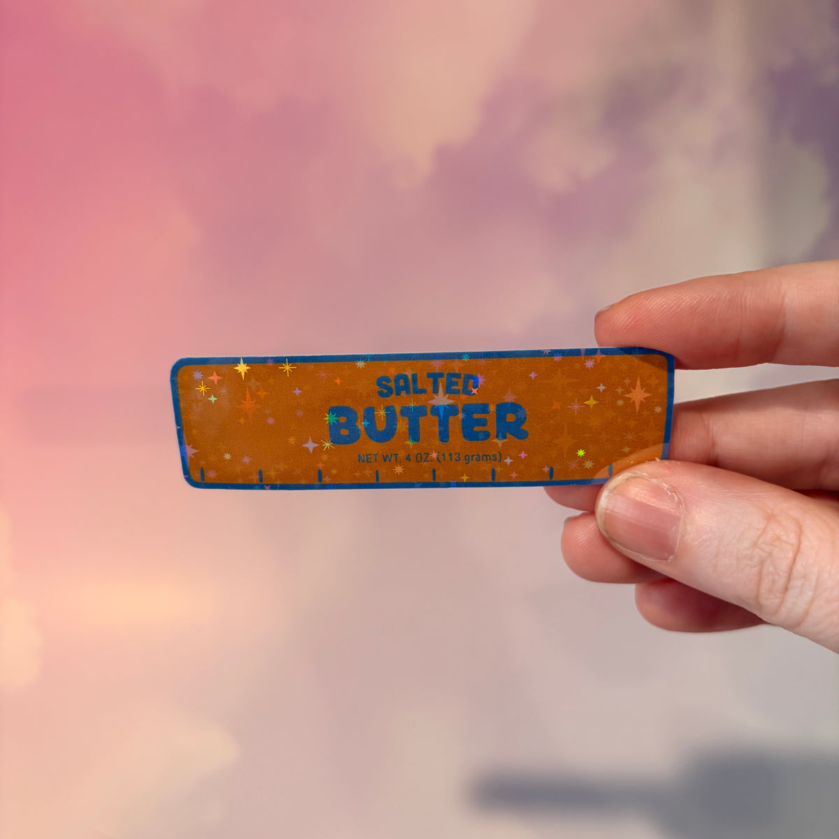 Holographic vinyl sticker shaped like a classic stick of butter, measuring approximately 3 inches long by 1 inch tall. Colorful reflective finish adds a playful touch to laptops, notebooks, tumblers, and sticker collections.