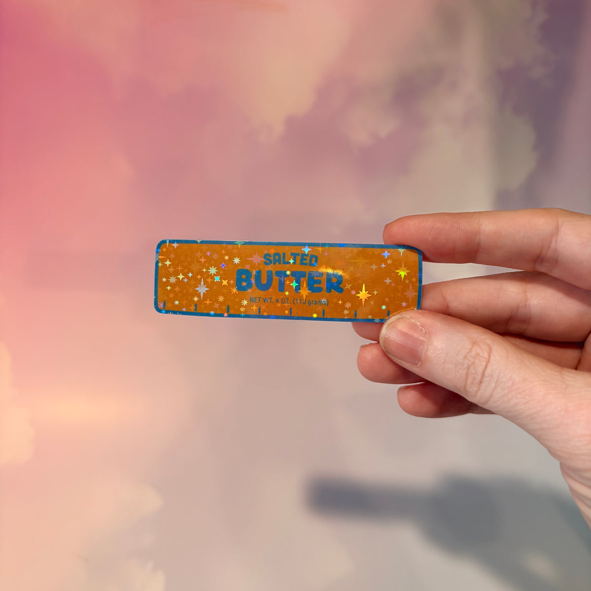 Holographic vinyl sticker shaped like a classic stick of butter, measuring approximately 3 inches long by 1 inch tall. Colorful reflective finish adds a playful touch to laptops, notebooks, tumblers, and sticker collections.
