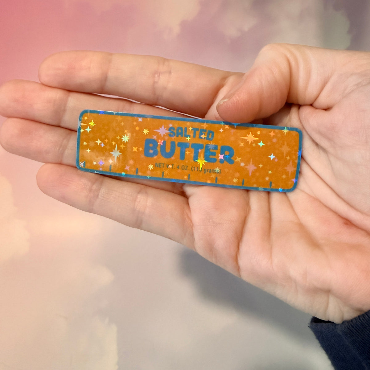 Holographic vinyl sticker shaped like a classic stick of butter, measuring approximately 3 inches long by 1 inch tall. Colorful reflective finish adds a playful touch to laptops, notebooks, tumblers, and sticker collections.