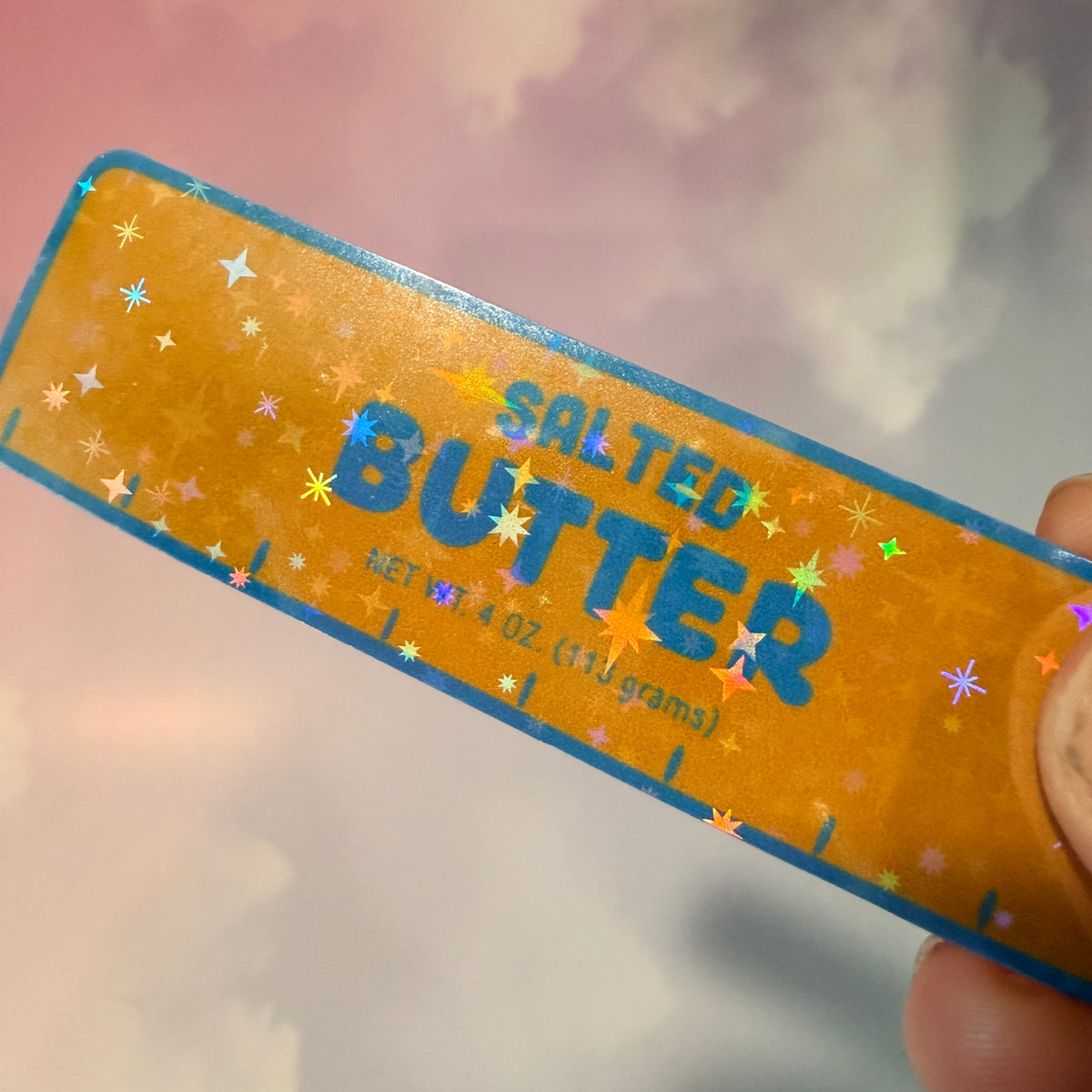 Holographic vinyl sticker shaped like a classic stick of butter, measuring approximately 3 inches long by 1 inch tall. Colorful reflective finish adds a playful touch to laptops, notebooks, tumblers, and sticker collections.